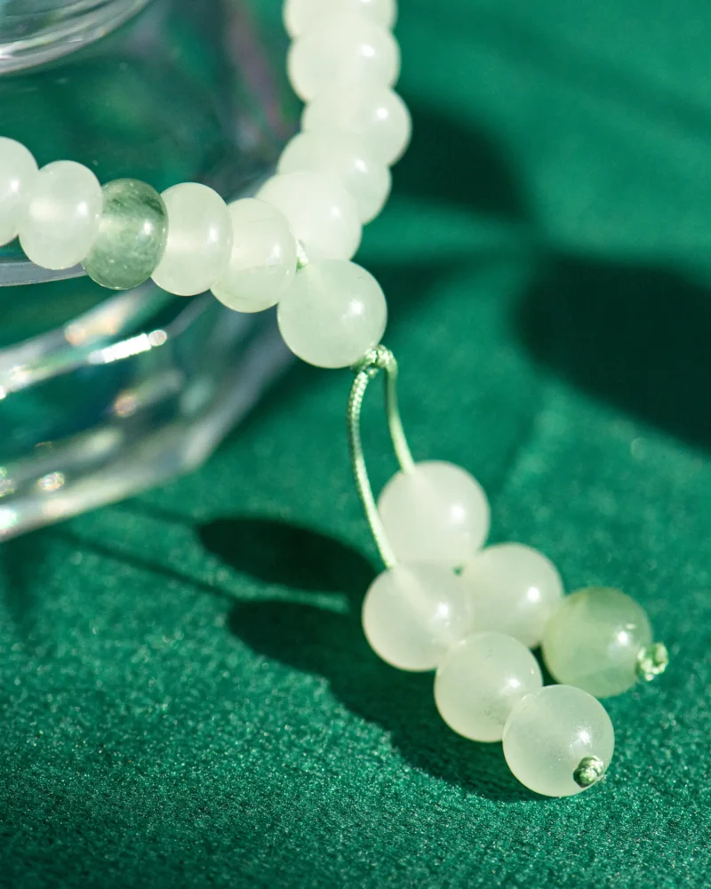 A close-up of the White and Light Green Quartzite Jade Beaded Bracelet with ring detail and twin drops, featuring tassels, resting on a green fabric surface with part of a glass object visible in the corner.