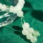A close-up of a white jade beaded bracelet with green accents, resting on a green fabric surface. The bracelet features a tassel with additional jade beads.