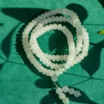 A string of round, translucent white beads is coiled on a textured green surface, with shadows of leaves and the beads creating patterns around it.