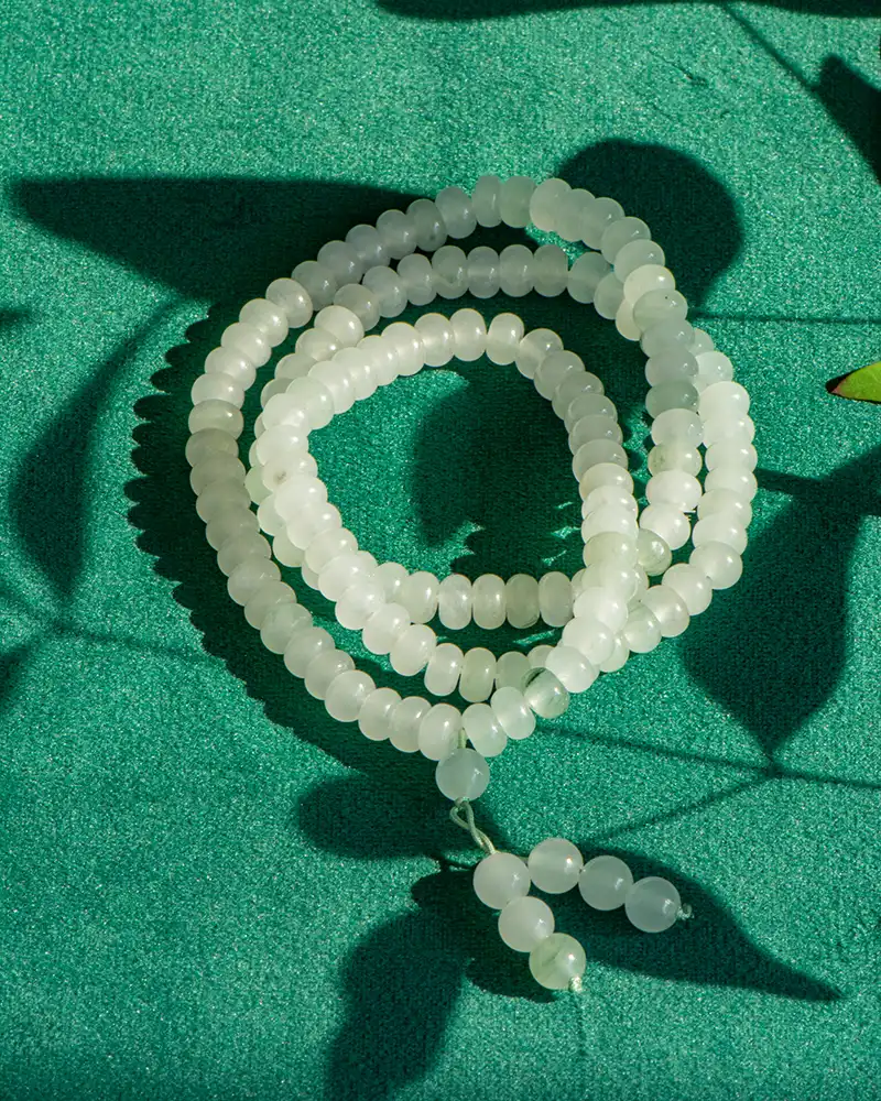 LYU_4871 A string of round, translucent white beads is coiled on a textured green surface, with shadows of leaves and the beads creating patterns around it.