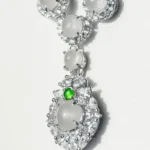 A close-up of a silver necklace with round, translucent gemstones surrounded by small clear stones and a single small green gem accent on the pendant, displayed on a light background.