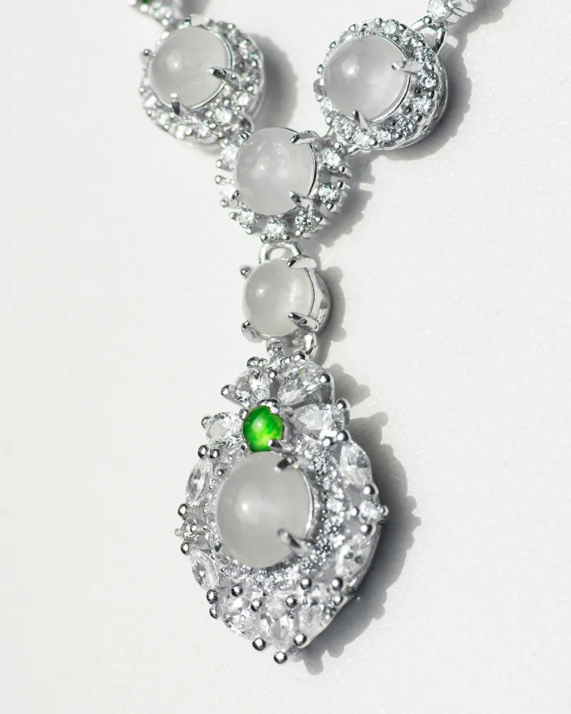 LYU_5011 A close-up of a silver necklace with round, translucent gemstones surrounded by small clear stones and a single small green gem accent on the pendant, displayed on a light background.