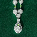 A silver necklace with round, translucent gemstones and small green beads, featuring an intricate, large pendant, displayed on a textured dark green background.