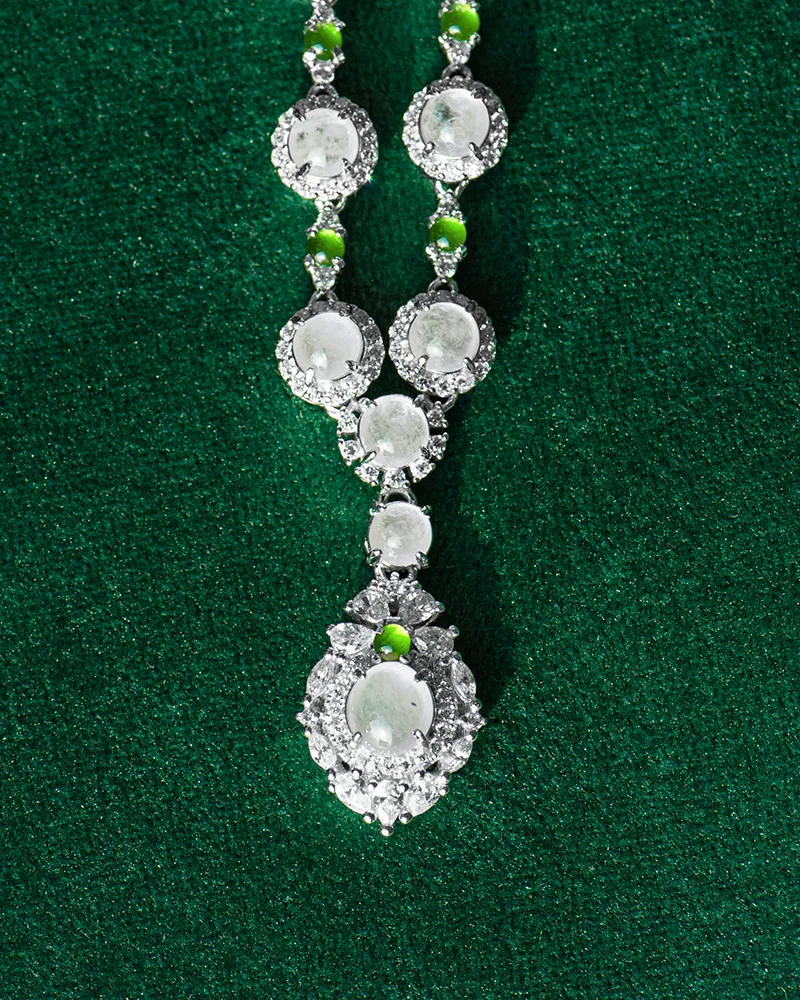 LYU_5015 A silver necklace with round, translucent gemstones and small green beads, featuring an intricate, large pendant, displayed on a textured dark green background.