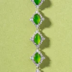 A bracelet with five oval green gemstones, each set in a diamond-shaped silver frame adorned with small clear stones, displayed on a light green background.