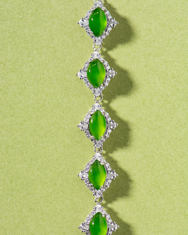LYU_5021 A bracelet with five oval green gemstones, each set in a diamond-shaped silver frame adorned with small clear stones, displayed on a light green background.