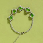 A silver bracelet with four marquise-shaped green stones, each set in a diamond-shaped cluster of clear crystals, displayed on a light green background. The bracelet has an adjustable chain with a small heart charm.