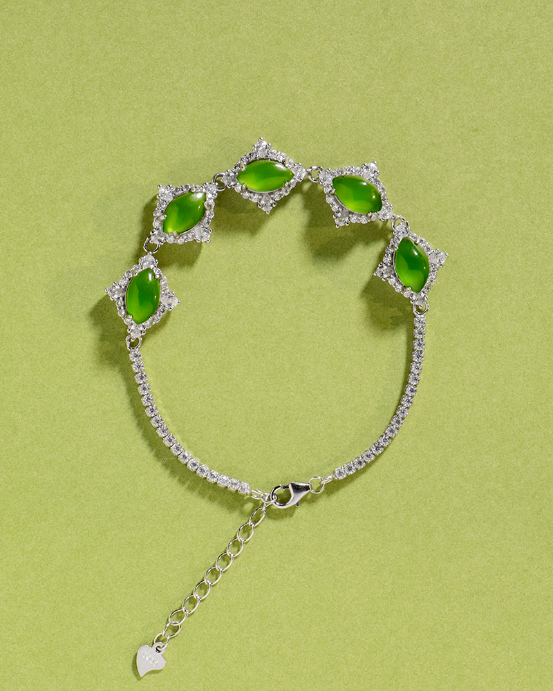 LYU_5025 A silver bracelet with four marquise-shaped green stones, each set in a diamond-shaped cluster of clear crystals, displayed on a light green background. The bracelet has an adjustable chain with a small heart charm.