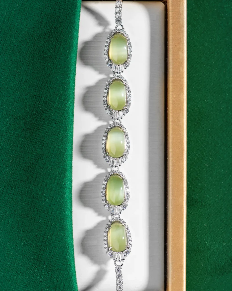 Presenting the Grayish-White Nephrite Jade Bracelet — Oval Cabochon Links: this elegant bracelet features five oval grayish-white nephrite jade cabochons, each accented with a halo of sparkling clear crystals. Shown resting on a white cushion against a green fabric backdrop, this exquisite piece highlights the refined beauty and timeless appeal characteristic of Val’s cabochon jewelry collections.