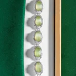 A silver bracelet with five oval green gemstones, each surrounded by small clear crystals, displayed in a jewelry box with green and white lining.