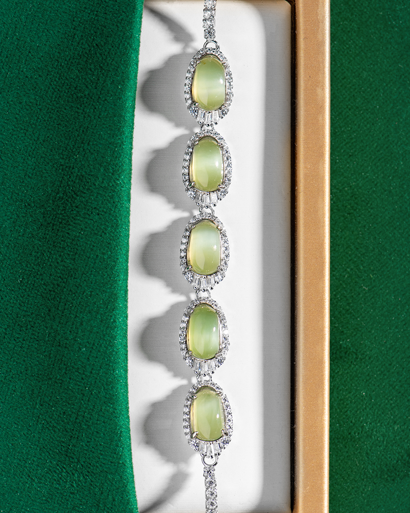 A silver bracelet with five oval green gemstones, each surrounded by small clear crystals, displayed in a jewelry box with green and white lining.