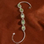 A silver bracelet with five oval green gemstones, each surrounded by small clear stones, displayed on a soft brown fabric background.