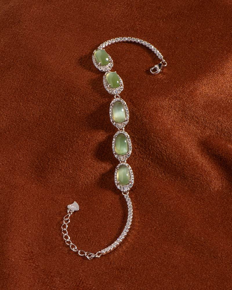 A silver bracelet with five oval green gemstones, each surrounded by small clear stones, displayed on a soft brown fabric background.