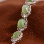 A close-up of a silver bracelet with four oval green gemstones, each surrounded by small clear crystals, displayed on a soft brown fabric background.