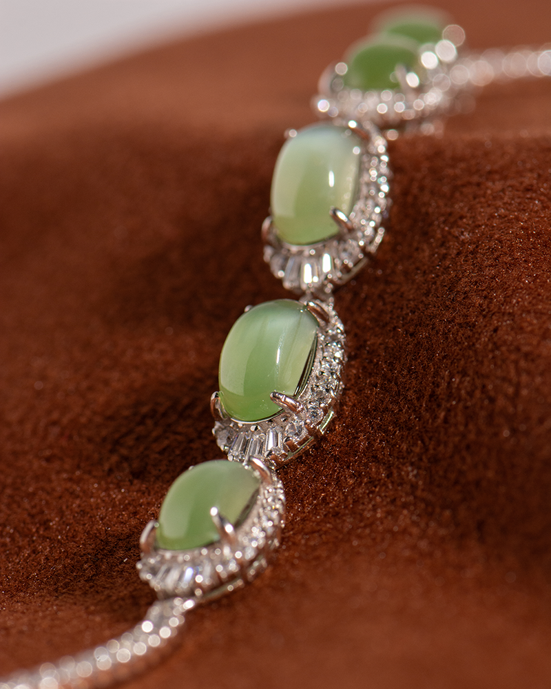 A close-up of a silver bracelet with four oval green gemstones, each surrounded by small clear crystals, displayed on a soft brown fabric background.