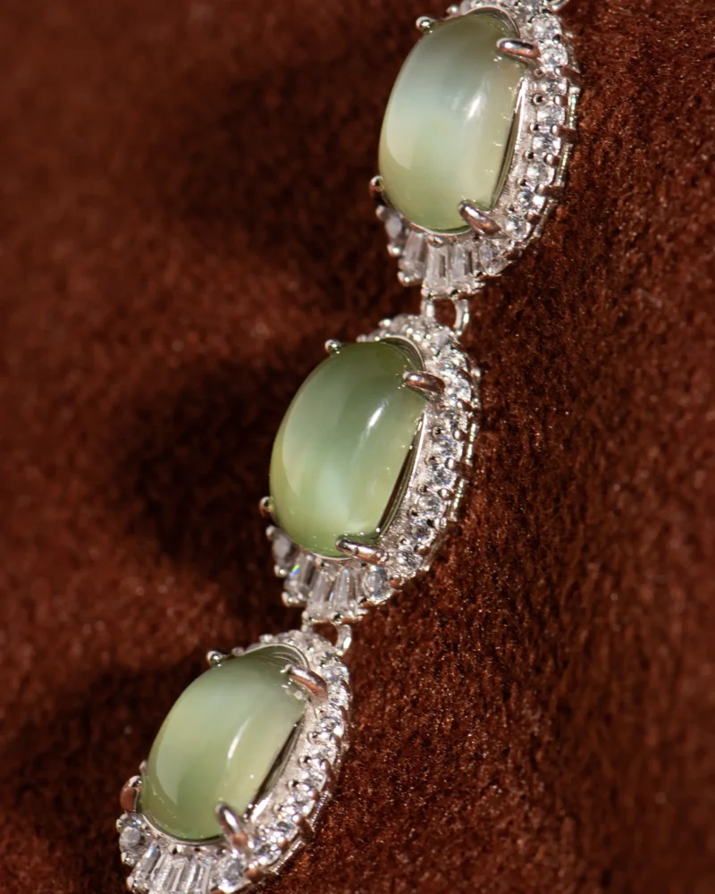 Close-up of the Grayish-White Nephrite Jade Bracelet — Oval Cabochon Links, showcasing three oval green nephrite cabochons bordered by small clear stones, arranged on a textured brown surface.