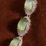 Close-up of a piece of jewelry featuring three oval green gemstones, each set in a silver, diamond-encrusted halo, displayed against a brown textured background.