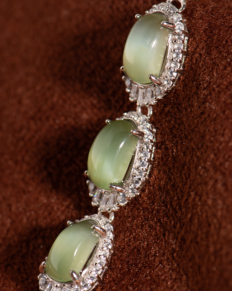 Close-up of a piece of jewelry featuring three oval green gemstones, each set in a silver, diamond-encrusted halo, displayed against a brown textured background.
