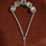 A silver bracelet with five pale green gemstones, each framed by sparkling stones, displayed on a brown textured surface. The bracelet features two chains joining at a clasp with an adjustable extender.