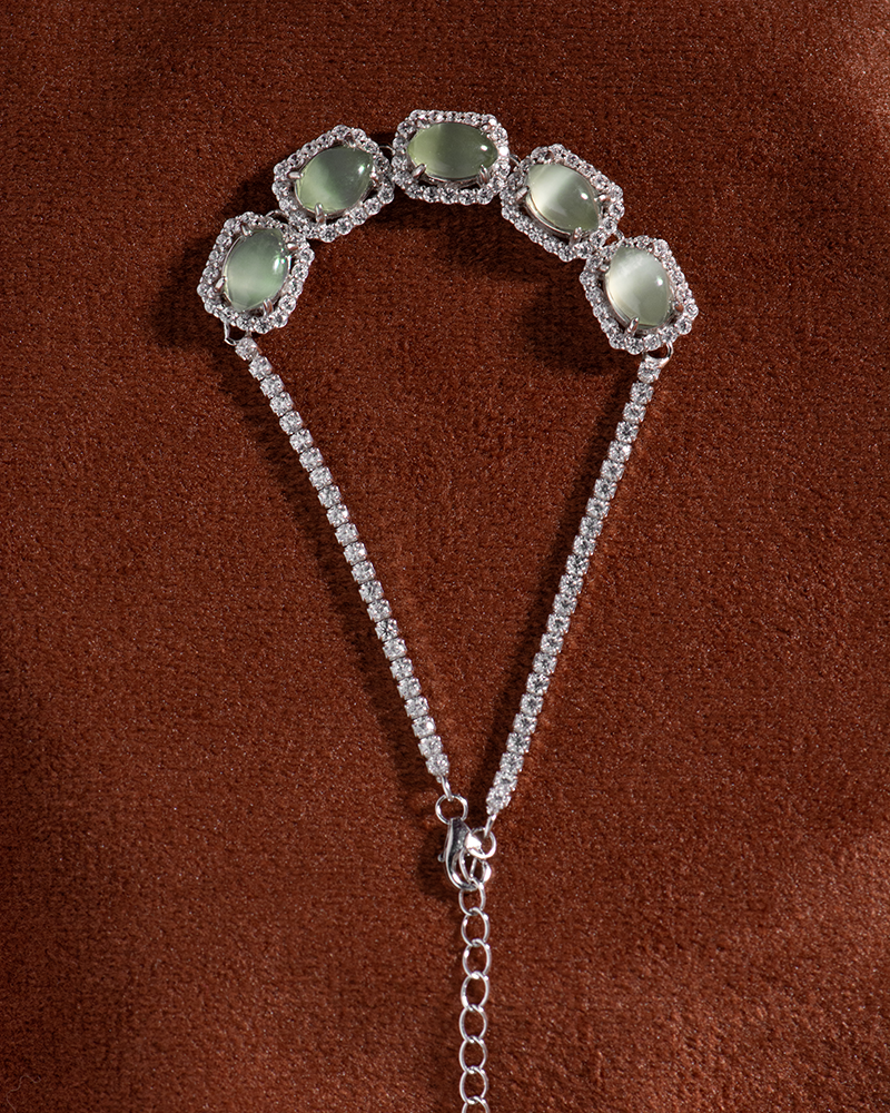 LYU_5037 A silver bracelet with five pale green gemstones, each framed by sparkling stones, displayed on a brown textured surface. The bracelet features two chains joining at a clasp with an adjustable extender.