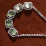 A diamond necklace featuring five oval green gemstones, each surrounded by a halo of diamonds, displayed on a rich brown textured background.