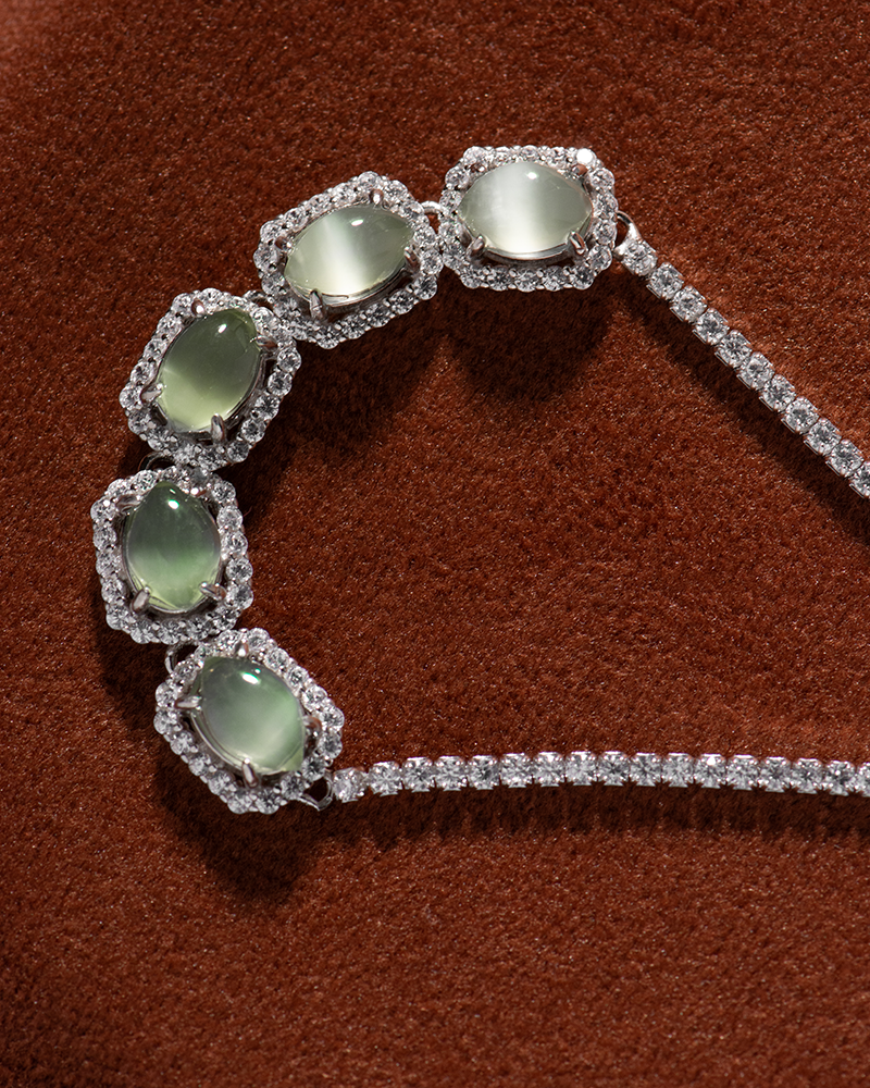 LYU_5038 A diamond necklace featuring five oval green gemstones, each surrounded by a halo of diamonds, displayed on a rich brown textured background.