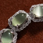 Close-up of three oval green gemstones, each surrounded by a halo of small, sparkling clear stones, set in silver metal, displayed on a textured brown background.