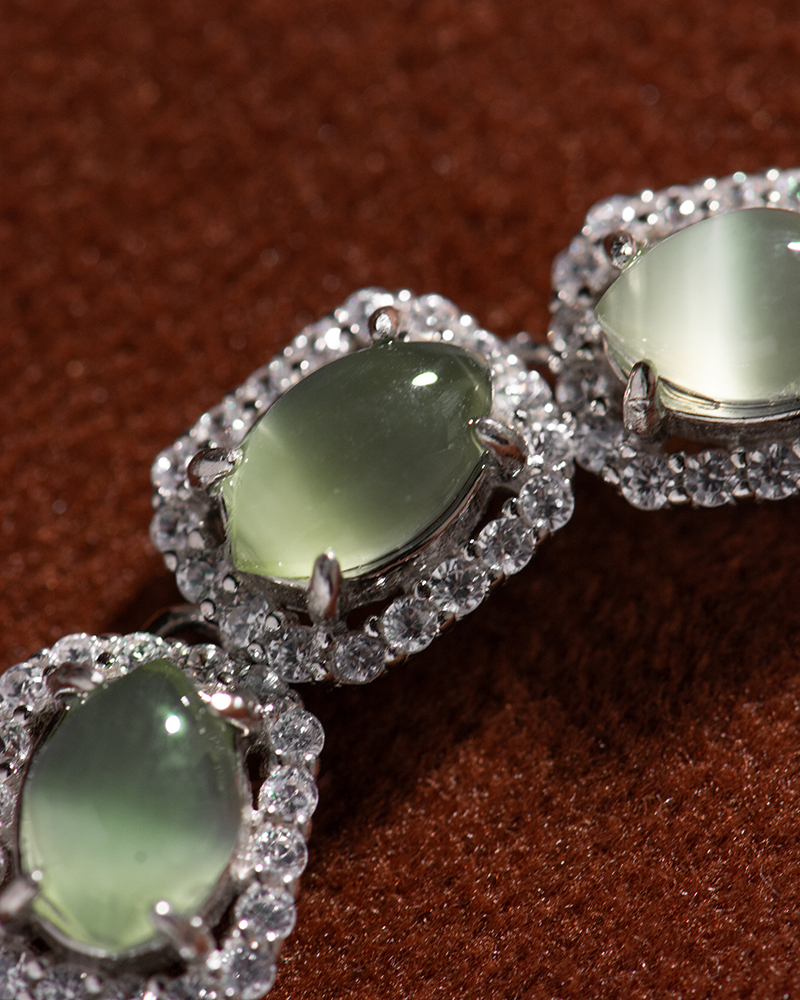 LYU_5039 Close-up of three oval green gemstones, each surrounded by a halo of small, sparkling clear stones, set in silver metal, displayed on a textured brown background.