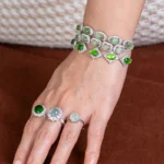 A hand with long, manicured nails is wearing two gemstone rings and two matching bracelets adorned with green and white stones, set in silver, resting on a brown surface.