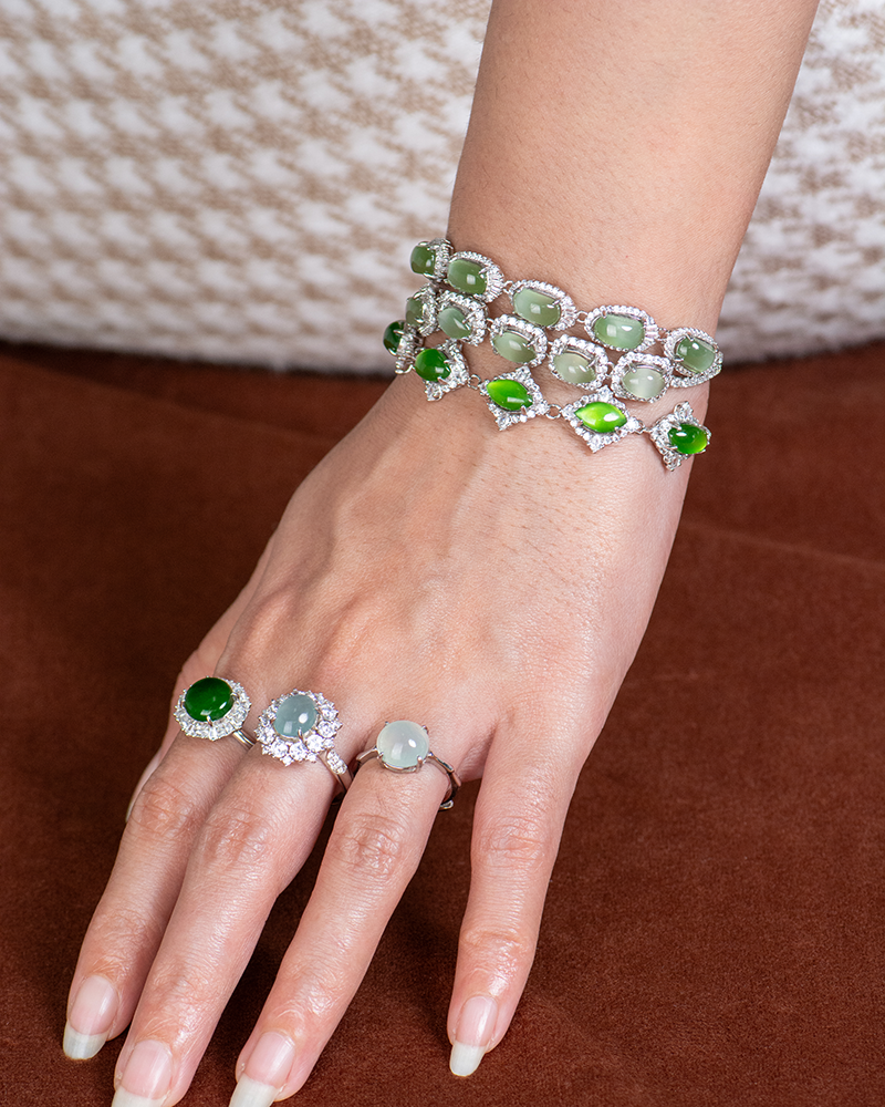 A hand with long, manicured nails is wearing two gemstone rings and two matching bracelets adorned with green and white stones, set in silver, resting on a brown surface.