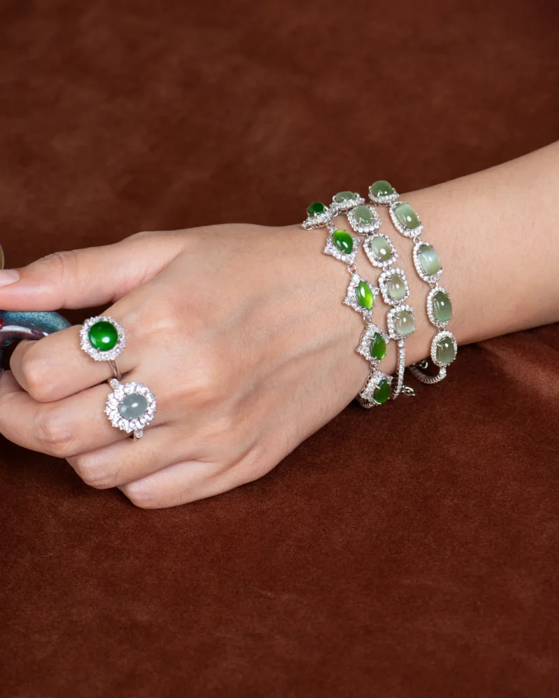 A hand resting on a brown surface is adorned with four Green Nephrite Jade Bracelets — Marquise Cabochon Link Line, along with two gemstone rings—one green and one clear—all set in silver with diamond accents. This elegant display highlights the distinctive marquise cabochon design of the jade bracelets, lending a unique touch to the overall look.