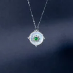 A silver necklace with a round, white pendant featuring a green gemstone flower design at the center, displayed against a dark blue background.