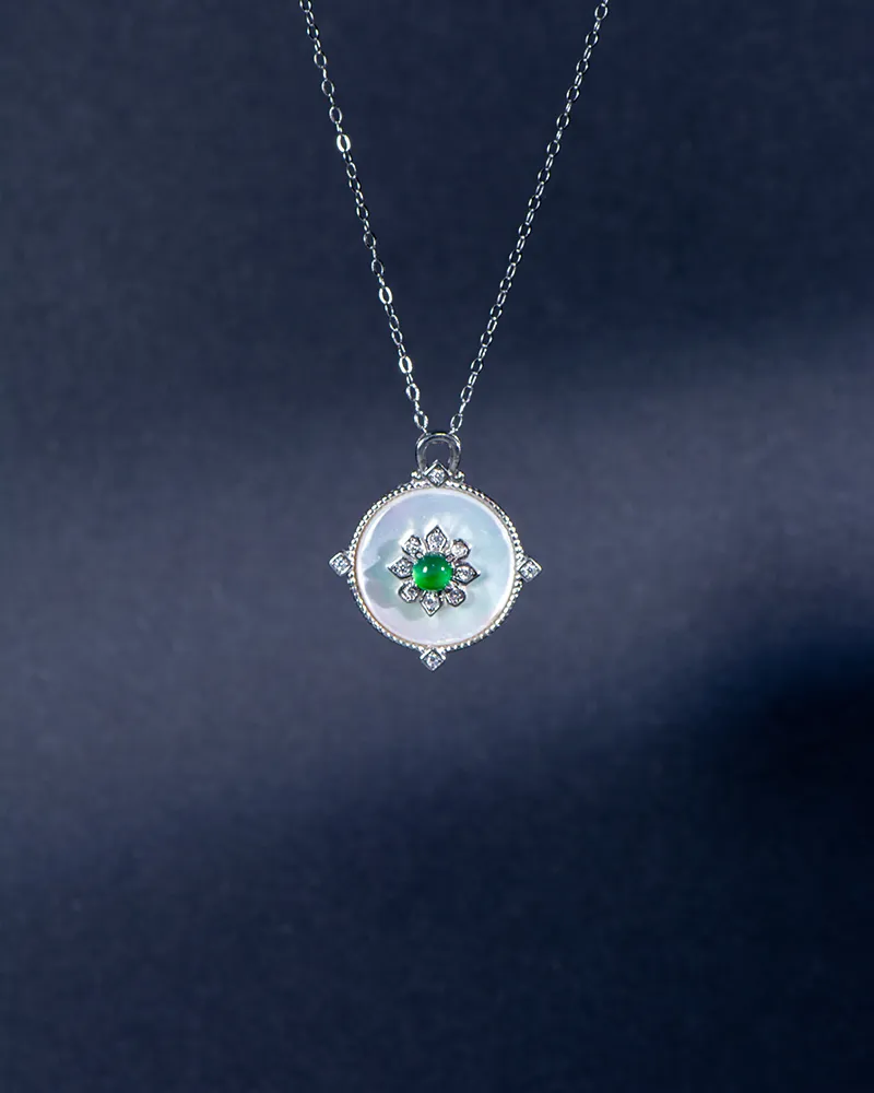 A silver necklace with a round, white pendant featuring a green gemstone flower design at the center, displayed against a dark blue background.
