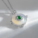 A silver necklace with a round, iridescent pendant featuring a central green gemstone surrounded by a floral arrangement of small diamonds, resting on a light surface.