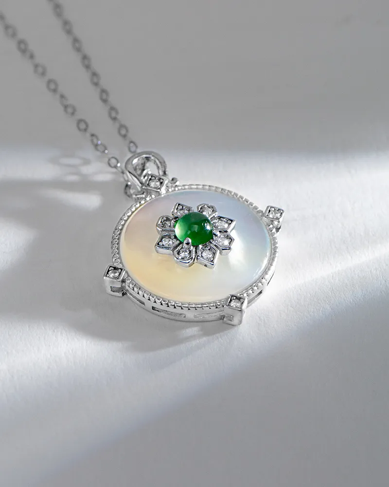 A silver necklace with a round, iridescent pendant featuring a central green gemstone surrounded by a floral arrangement of small diamonds, resting on a light surface.