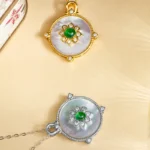 Two round pendants with floral designs, one in gold and the other in silver, each featuring a central green stone, pearl-like background, and diamond accents, displayed on a beige surface.