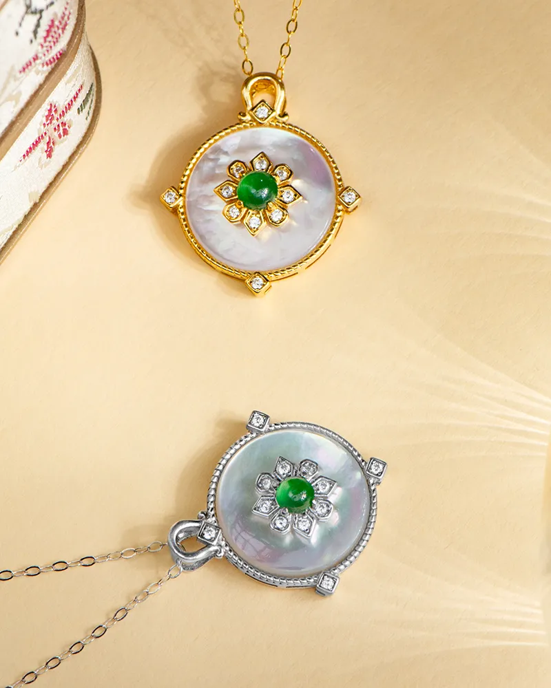 LYU_5458 Two round pendants with floral designs, one in gold and the other in silver, each featuring a central green stone, pearl-like background, and diamond accents, displayed on a beige surface.