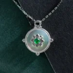 A round silver pendant necklace featuring a white iridescent center with a floral design, set with a small green gemstone in the middle, displayed on a silver chain against dark green and black backgrounds.