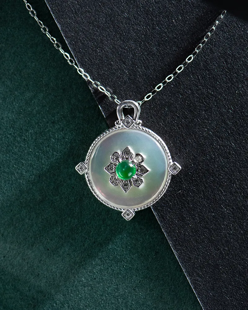 A round silver pendant necklace featuring a white iridescent center with a floral design, set with a small green gemstone in the middle, displayed on a silver chain against dark green and black backgrounds.