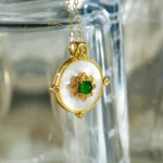 A gold necklace with a round pendant, featuring a white center, gold floral design, and a green gemstone in the middle, hanging on the edge of a clear glass surface.