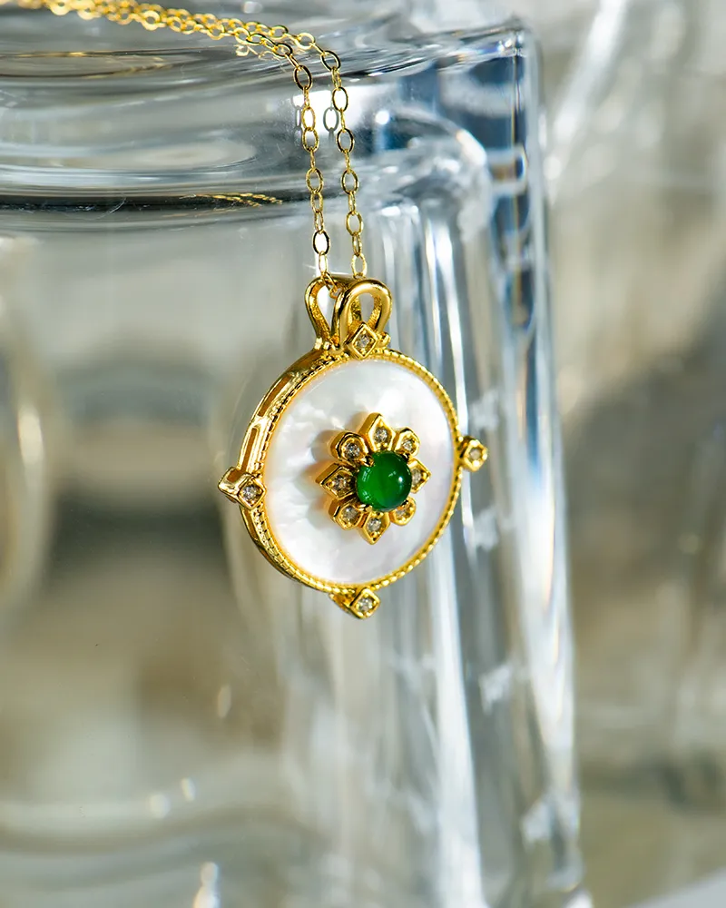 LYU_5491 A gold necklace with a round pendant, featuring a white center, gold floral design, and a green gemstone in the middle, hanging on the edge of a clear glass surface.