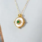 A gold pendant necklace featuring a round white mother-of-pearl disk, adorned with a central green gemstone and small gold details, displayed against a light gray background.