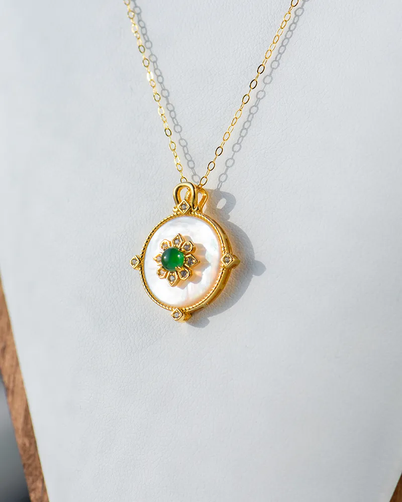 LYU_5498 A gold pendant necklace featuring a round white mother-of-pearl disk, adorned with a central green gemstone and small gold details, displayed against a light gray background.