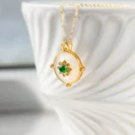 A gold necklace with a round pendant featuring a green gemstone and small clear stones is displayed hanging over the rim of a white, textured ceramic bowl.