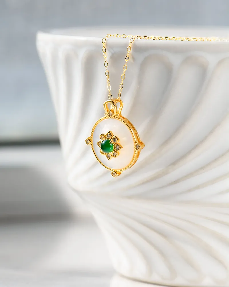 LYU_5504 A gold necklace with a round pendant featuring a green gemstone and small clear stones is displayed hanging over the rim of a white, textured ceramic bowl.