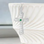 A silver cross pendant with intricate detailing and a green gemstone at the center hangs in front of a white, textured ceramic bowl.
