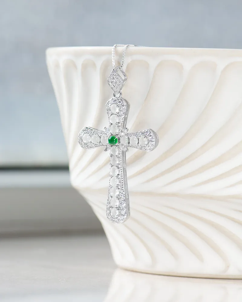 A silver cross pendant with intricate detailing and a green gemstone at the center hangs in front of a white, textured ceramic bowl.