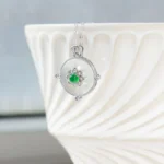 A silver pendant necklace with a circular white gemstone and a green stone at the center, accented by small clear stones, hangs over the edge of an elegant, textured white ceramic bowl.