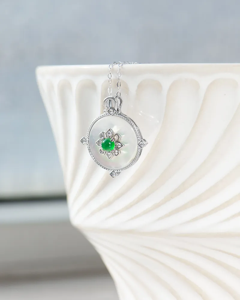 A silver pendant necklace with a circular white gemstone and a green stone at the center, accented by small clear stones, hangs over the edge of an elegant, textured white ceramic bowl.
