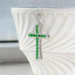 A silver cross pendant with green gemstones and small clear stones hangs against a white, textured ceramic bowl. The background is softly blurred, highlighting the jewelry.